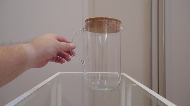 A clear glass pitcher with a cork lid is carefully pushed by a hand across a glass shelf. The glass base creates a brief scraping and grinding friction sound on the hard surface.