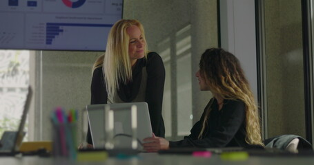 Blonde businesswoman leaning over desk to explain point to seated colleague during close collaboration in office with data chart on display