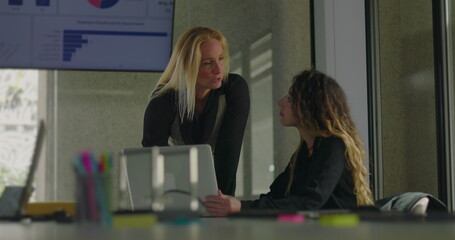 Blonde businesswoman leaning over desk to explain point to seated colleague during close collaboration in office with data chart on display
