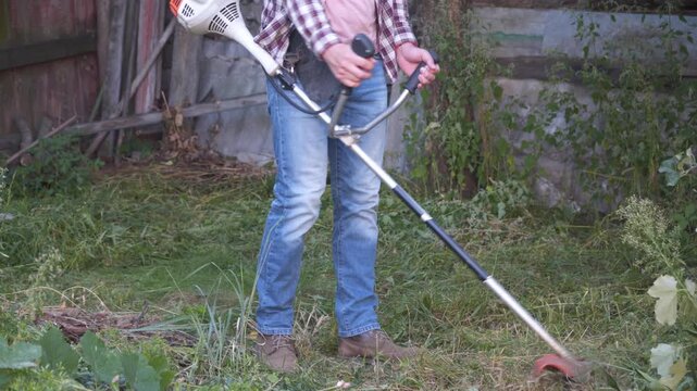 Gardener works in yard of rural house with lawn mower. Male farmer cutting wild grass with an electric scythe at the farm. Young male worker cutting grass weeds, using trimmer. Gasoline brush cutter.