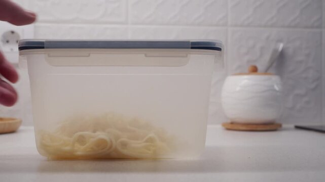 A person's hand places a plastic food storage container with cooked noodles onto a white kitchen counter. The plastic container makes a dull thud as it is set down.
