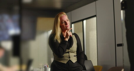 Fototapeta premium Blonde businesswoman with preoccupied expression gazing into distance in office, reflecting on future while team works in background near conference table
