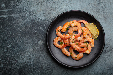 Cooked shrimps prawns arranged on a black plate with lemon wedges and chopped parsley, elegant top view seafood appetizer on dark background space for text