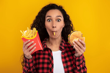A joyful lady is having a fun time eating a burger and french fries, holding a piece of potato in her mouth. The vibrant yellow background adds to the playful mood of this fast food moment.