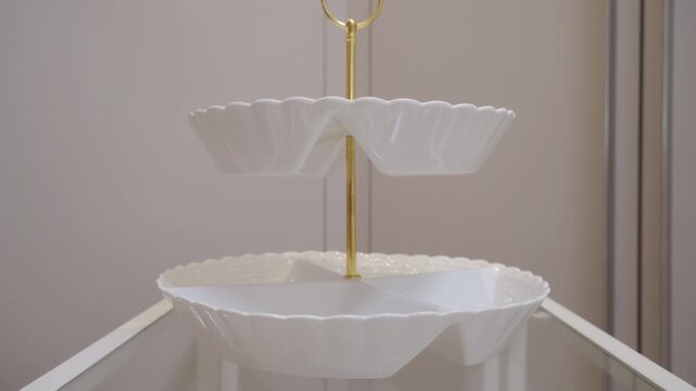 White glossy ceramic and gold metal tiered serving dish is set down on a glass surface. A light, delicate clink rings out.