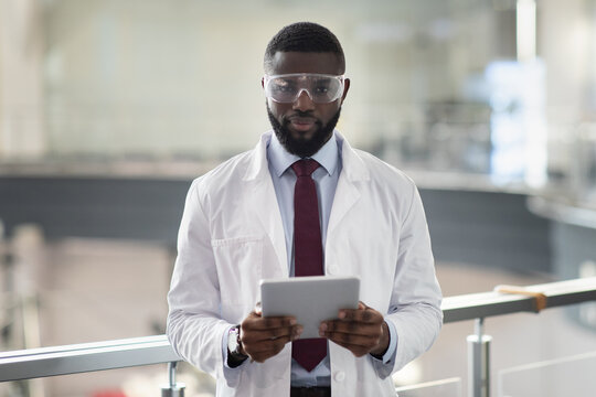 Cheerful young black man in workwear and protective glasses medical scientist using digital tablet, analyzing data, posing at workplace, panorama with copy space. Jobs and occupations concept