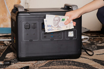 A girl holds hryvnias near a portable power station in an apartment connected to provide backup power during a power outage