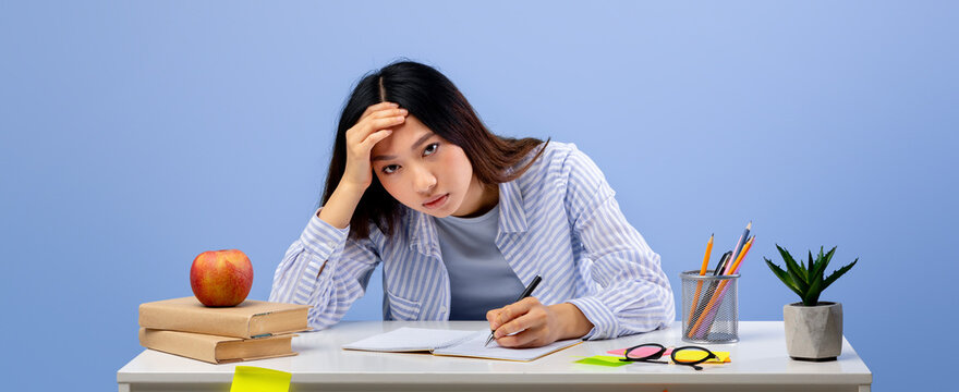 A student is sitting at a desk with books and an apple, looking stressed while writing in a notebook. She appears to be deep in thought and struggling with her work.
