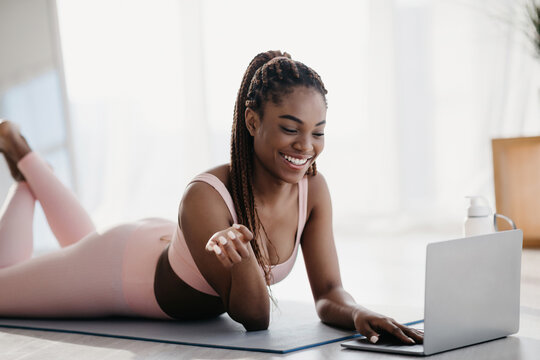 A joyful African American woman in a sports outfit lies on her fitness mat, connecting with her personal trainer online using her laptop. She is ready for a yoga class at home.