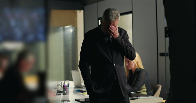 Businessman deeply stressed with hands over face while standing in meeting room as female colleague works in background on laptop during corporate crisis