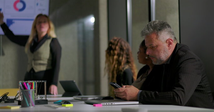 Businessman discreetly checking phone during office meeting as female colleague presents, casual modern workplace dynamics with multitasking and quiet team engagement
