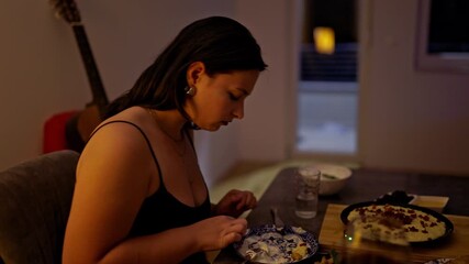 A young woman enjoys a solitary dinner, savoring her meal in the cozy ambiance of her home during a peaceful evening. This intimate moment captures personal comfort and relaxation.