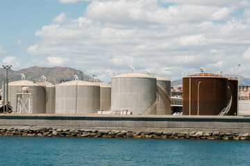 Obraz premium Industrial storage tanks by the sea with mountain backdrop and cloudy sky