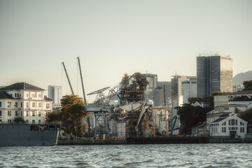Huge offshore construction vessel with towering cranes and complex machinery docked at urban harbor for maintenance; framed by old white warehouses, modern skyscraper skyline and rippling water