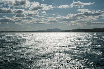 Obraz premium Shimmering sea under partly cloudy skies with distant mountain view