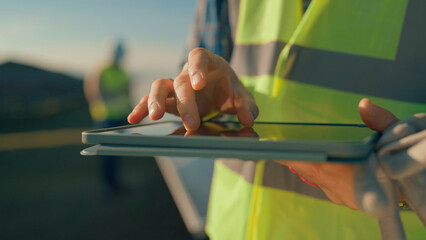 Worker uses tablet for data entry in construction site during sunset with colleague in background