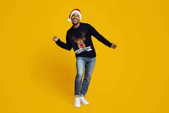 A man smiles and dances cheerfully while wearing a Christmas-themed sweater and a Santa hat. The bright yellow background adds to the festive atmosphere, making the scene lively. - Powered by Adobe