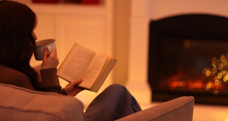 Woman with cocoa drink reading book in armchair near fireplace at home, space for text