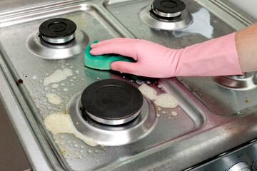 Cleaning a dirty gas stove with sponge and detergent. Hand in rubber glove scrubbing grease from stainless steel surface.