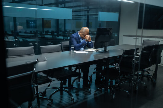 Mature African American businessman sitting with head bowed in a quiet meeting room, showing exhaustion or deep concentration. Corporate stress and workplace pressure reflected clearly