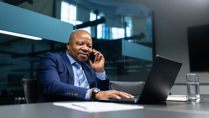 African American businessman speaking on cellphone while working on laptop in modern office. Confident mature executive managing tasks with ease and focus