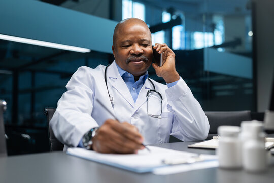 Mature African American doctor talking on the phone while writing notes at his desk. Professional physician managing patient records and medical tasks in a modern clinic office