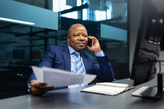 Mature African American businessman on a phone call while holding documents in modern office. Confident executive discussing work matters with steady, professional focus