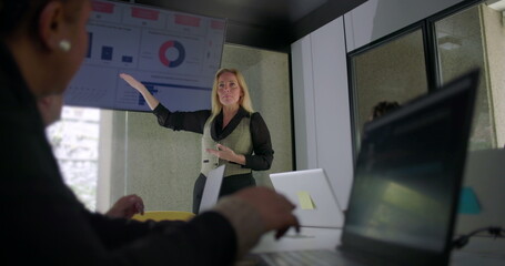 Businesswoman explaining data during meeting as team listens attentively in modern office with laptops and digital charts on display screen