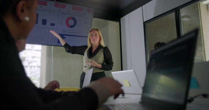 Businesswoman explaining data during meeting as team listens attentively in modern office with laptops and digital charts on display screen - Powered by Adobe