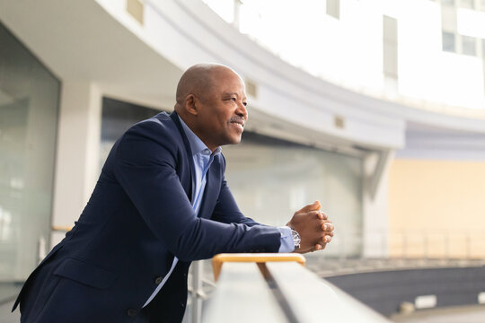 Mature African American businessman leaning on railing with a thoughtful smile, taking a quiet moment to reflect in a bright modern building. Calm executive enjoying a peaceful break