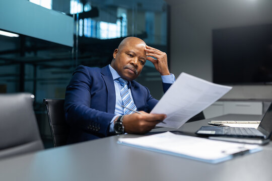 Serious mature African American businessman analyzing documents in modern office. Focused corporate leader reviewing paperwork with a thoughtful, concentrated expression - Powered by Adobe
