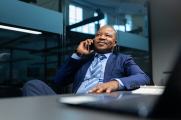 Relaxed mature African American businessman leaning back while talking on cellphone in modern office. Confident executive maintaining a calm and positive business conversation