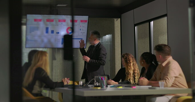 Business executive presenting analytics to diverse team in glass-walled office conference room during strategy meeting