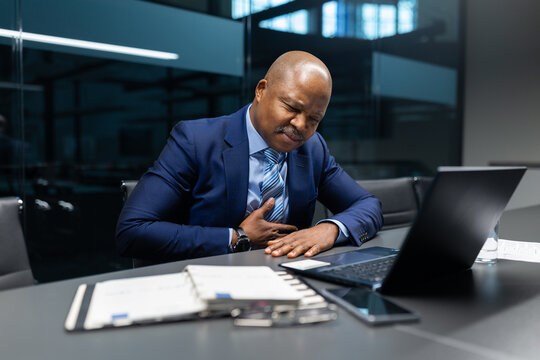 Mature african american businessman holding his chest while sitting at his office desk, showing sudden discomfort or stress during a busy workday in a modern corporate environment