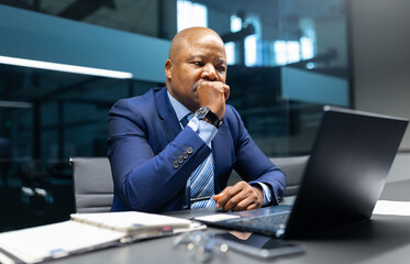 A mature African American businessman sitting at his desk, thinking seriously while looking at his laptop, reflecting problem-solving and strategic planning at work