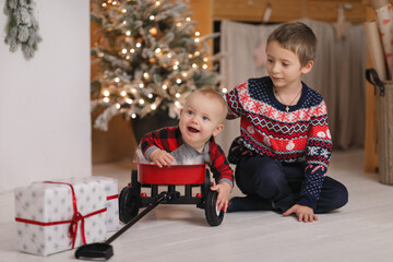Happy baby boy sitting in a red wagon toy, being watched by his older brother in a christmas sweater near a glowing holiday tree and gifts