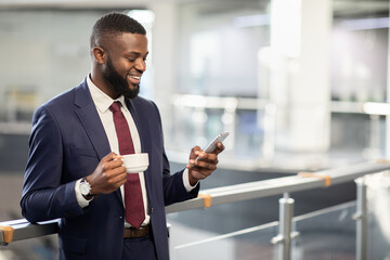 Cheerful young black bearded man manager standing at office building corridor, drinking coffee,...