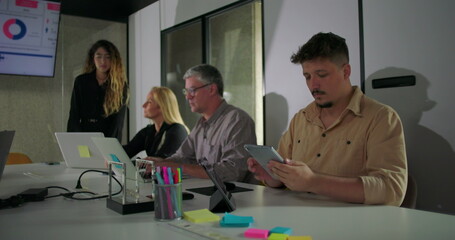 Business meeting with diverse team focused on digital presentation, as male employee on right uses tablet while others engage with charts and laptops around conference table