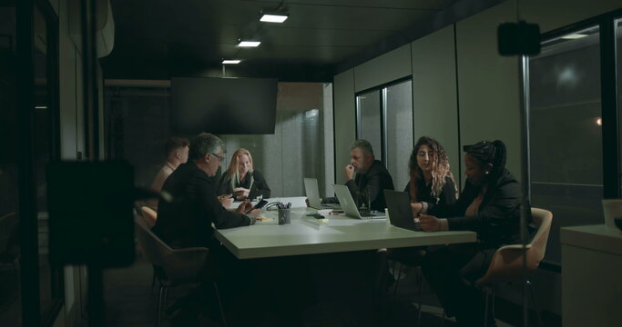Business professionals in formal meeting room working on laptops and tablets during evening corporate discussion around white conference table inside glass office