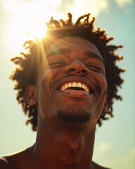 Young man with dark skin and curly hair laughing joyfully with eyes closed, showing bright white teeth and a wide, happy smile, basking in warm sunlight.