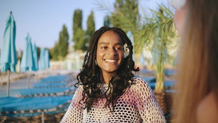 Multiracial smiling friends with curly hair wearing a colorful cover-up enjoying time outdoors near a sea on a sunny day surrounded by palm trees and lounge chairs