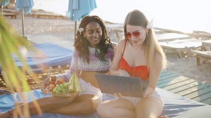 Multiracial female Friends enjoying fruit and browsing on a laptop at a sunny beach during the...