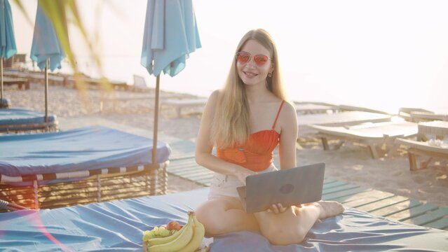 Young woman enjoying a sunny beach day while working on a laptop, surrounded by fruits and lounge chairs, capturing a relaxed summer vibe