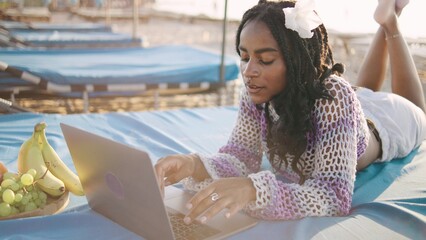 African American woman working on a laptop while relaxing on a beach lounge during sunset with fresh fruits nearby