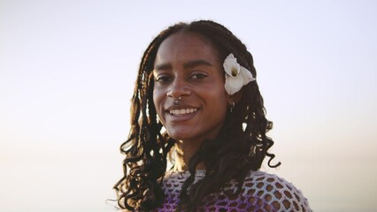 Portrait of a young African American woman smiling by the ocean at sunset with a flower in her hair, showcasing natural beauty and confidence in a coastal setting