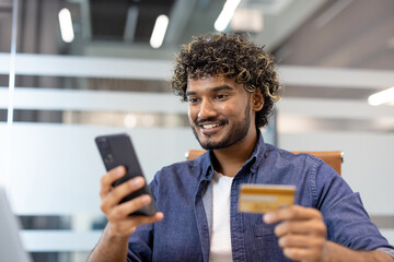 Indian smiling young man sitting at office desk, using mobile phone and holding credit card