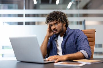 Upset young Indian man sitting at a desk in the office and looking at the laptop screen in frustration