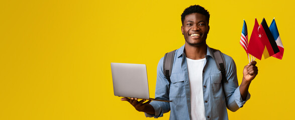 A young man stands smiling, holding multiple flags in one hand and a laptop in the other. Bright...