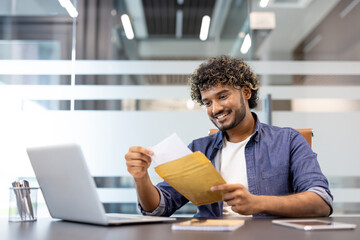A young Indian man sits at a desk in an office and smiles as he reads a letter he received in an...