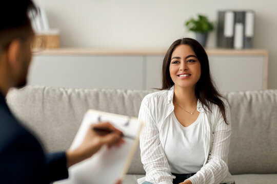 Happy Arab female client engages in a successful psychotherapy session with her psychologist. She smiles while discussing her emotional challenges, creating a positive atmosphere at the clinic.